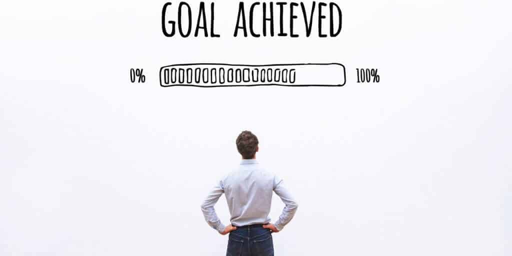 man in a room looking up at black lettering that reads 'Goal Achieved' alongside a fictitious loading bar from 0% to 100%